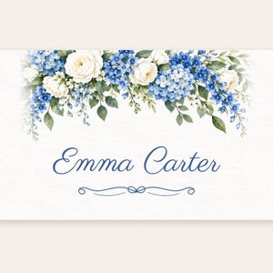 May include: A rectangular white card featuring a watercolour floral arrangement of blue hydrangeas, white roses, and green leaves along the top edge. The name Emma Carter is printed in blue script in the centre, with a decorative blue flourish below.