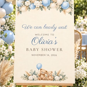 May include: A rectangular welcome sign for a baby shower featuring the text We can bearly wait, WELCOME TO Olivia's BABY SHOWER, and NOVEMBER 14, 2026. The design includes blue and cream balloons, white flowers, and two brown teddy bears.