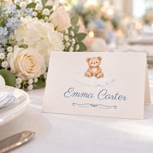May include: A white folded place card featuring a brown teddy bear illustration sitting on a light blue cloud. The name Emma Carter is printed in blue script below the bear, with a small blue decorative flourish underneath the text.