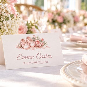 May include: A white folded place card featuring a watercolor illustration of pink baby shoes and roses at the top. The name Emma Carter is printed in a pink script font in the center, with a decorative flourish line underneath the name.
