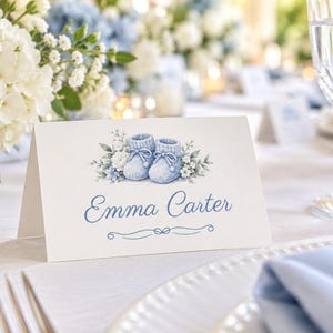 May include: A white folded place card featuring a watercolor illustration of light blue knitted baby booties surrounded by small white and blue flowers. The name Emma Carter is printed in a blue script font below the illustration.