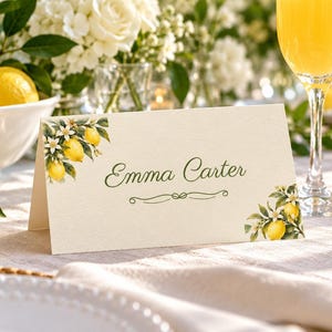 May include: A folded cream-colored place card featuring a printed illustration of yellow lemons and white blossoms in the top-left and bottom-right corners. The name Emma Carter is printed in dark green script in the center of the card.