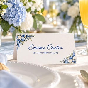 May include: A white folded place card featuring a blue floral watercolor design in the top left and bottom right corners. The name Emma Carter is printed in a blue script font in the center, with a decorative blue line element below it.