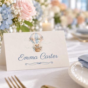 May include: A folded white place card featuring a watercolour illustration of a teddy bear in a hot air balloon with blue and pink accents. The name Emma Carter is printed in blue script below the illustration, with a decorative blue line.