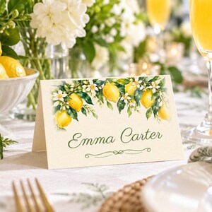 May include: A folded white place card featuring a watercolor illustration of yellow lemons, green leaves, and white blossoms along the top edge. The name Emma Carter is printed in a dark green script font in the center of the card.