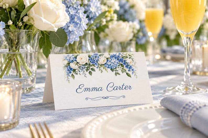 May include: A white folded place card featuring a watercolour floral illustration of blue hydrangeas and white roses across the top. The name Emma Carter is printed in a blue script font in the centre, with a decorative blue line underneath.
