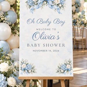 May include: A rectangular welcome sign on a wooden easel featuring a light blue watercolor background, white and blue floral arrangements, and a blue ribbon. The text reads: Oh Baby Boy, WELCOME TO, Olivia's, BABY SHOWER, NOVEMBER 14, 2026.