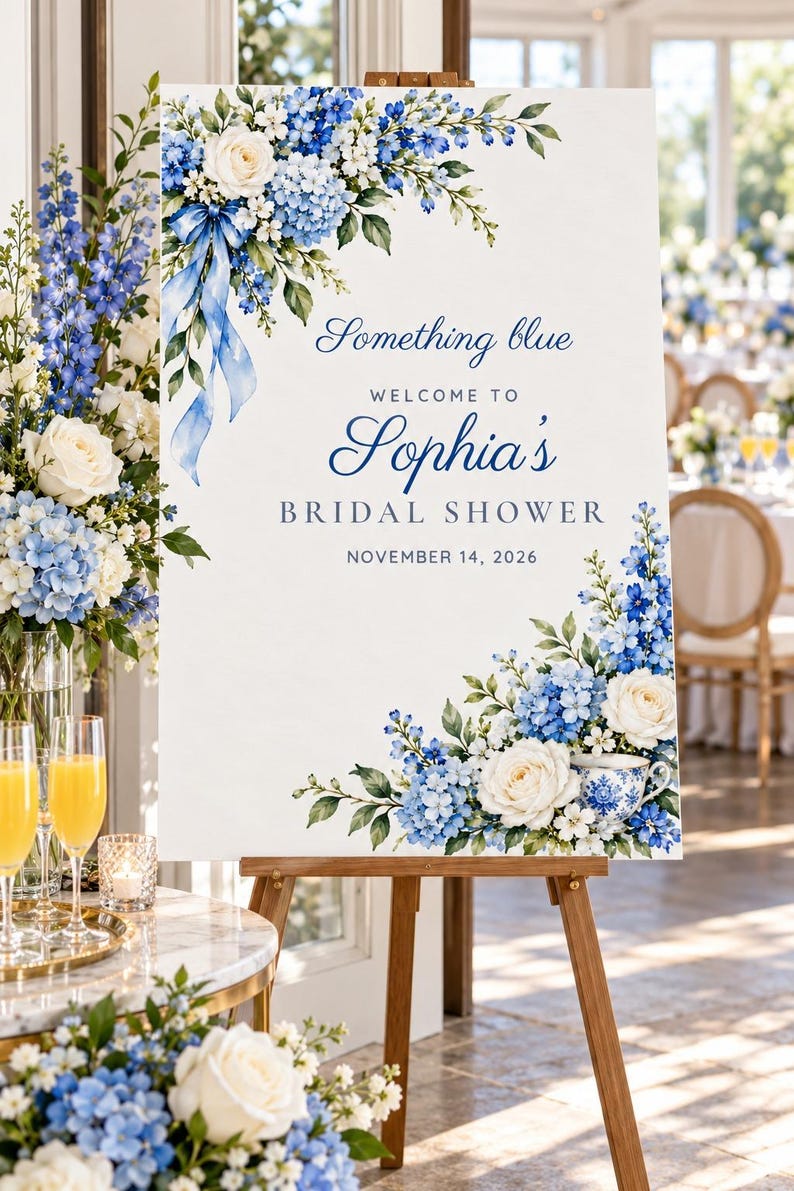May include: A white rectangular bridal shower welcome sign on a wooden easel, featuring blue and white floral arrangements, a blue ribbon, and a teacup. The text reads: Something blue, WELCOME TO Sophia's BRIDAL SHOWER, NOVEMBER 14, 2026.