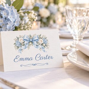May include: A white folded place card featuring a watercolor illustration of blue hydrangeas, white roses, and a light blue ribbon bow. The name Emma Carter is printed in blue script font below the floral design, with a small decorative flourish.