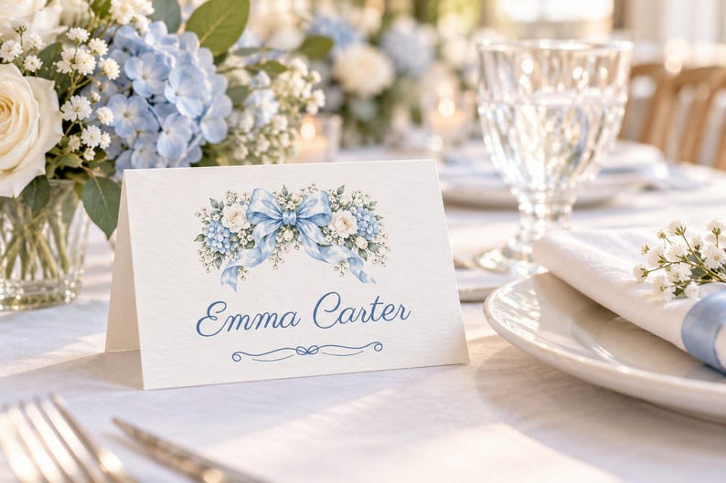 May include: A white folded place card featuring a printed illustration of blue and white flowers with a central blue ribbon bow. The name Emma Carter is printed in blue script below the floral design, with a small decorative flourish underneath.