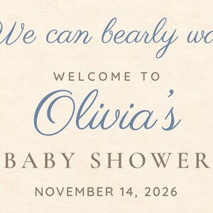 May include: A cream-colored sign with blue and brown text that reads: We can bearly wait, WELCOME TO, Olivia's, BABY SHOWER, NOVEMBER 14, 2026. The text uses a mix of elegant cursive script and clean, capitalized sans-serif typography.