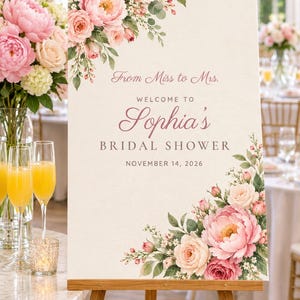 Op de afbeelding: Een rechthoekig welkomstbord op een houten ezel met roze en witte bloemstukken in de linker bovenhoek en rechter onderhoek. De tekst luidt From Miss to Mrs. WELCOME TO Sophia's BRIDAL SHOWER NOVEMBER 14, 2026.