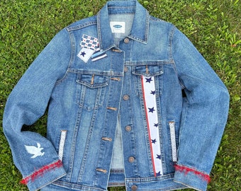May include: A light blue denim jacket with patriotic embellishments. Features a white and red stripe with blue stars, patches, and an eagle graphic. The cuffs have red fringe and white lace trim.