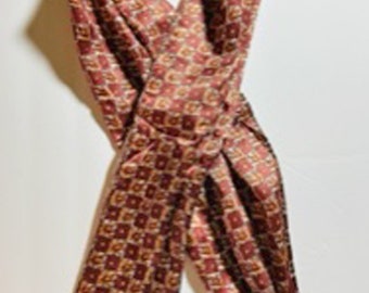 May include: A burgundy scarf with a repeating square pattern in shades of red, gold, and cream. The scarf has a decorative border with a floral design and fringe at the ends. The scarf is made of a soft, flowing material.