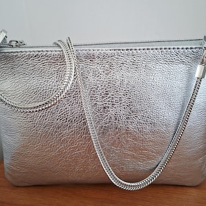 May include: Silver clutch bag with a textured finish and a silver chain strap. The rectangular bag has a zip closure and a small silver chain handle. The bag is on a wooden surface.