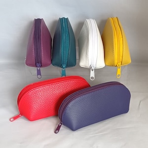 May include: Six different colored zippered pouches, all in a half-moon shape. The pouches are made of a textured leather-like material. The colors are purple, teal, white, yellow, red, and blue.