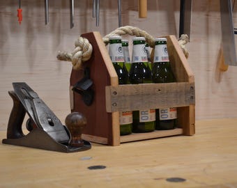 May include: A wooden beer caddy with a thick rope handle and a black metal bottle opener attached to the side, holding several green glass beer bottles. Next to the caddy sits a metal hand plane with a dark wooden handle and knob.