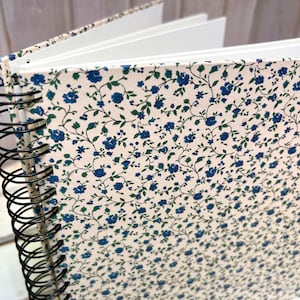 Può includere: Un quaderno a spirale color crema con un motivo floreale blu e verde sulla copertina.