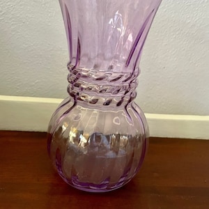 May include: A light purple glass vase with a round base and a twisted design around the middle.