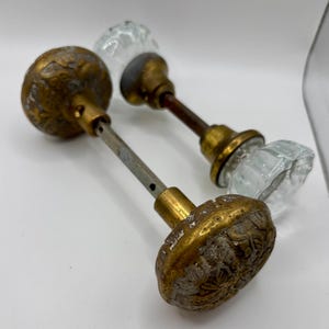 Puede incluir: Dos pomos de puerta con un eje metálico central. Uno presenta una cara redonda de latón texturizado con detalles florales y una parte posterior de cristal transparente. El otro tiene una cara de cristal transparente facetado con base de latón.