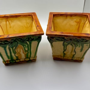 Peut inclure: Une paire de cache-pots carrés en céramique avec une base de couleur crème et une finition brillante. Les bords sont émaillés d'un brun ambré chaud, tandis que les côtés présentent des motifs de feuilles en relief et des coulures vertes.