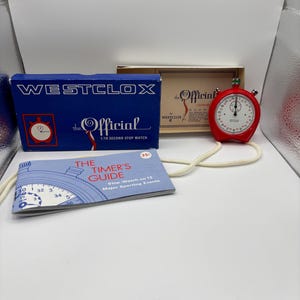 Può includere: Un cronometro Westclox rosso vintage con quadrante bianco e cordino color crema. Il cronometro è accanto a una scatola blu e una beige, entrambe con l'etichetta "Westclox The Official". È presente anche un piccolo libretto intitolato "The Timer's Guide".