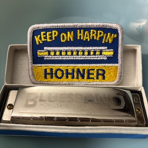 May include: A Hohner Bluesband harmonica in a blue and white box, with a patch that says "KEEP ON HARPIN'" and "HOHNER". The harmonica is silver with the word "BLUESBAND" engraved on it.