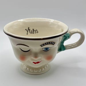 May include: Cream-colored ceramic mug with a winking face design, red lips, and rosy cheeks. The mug features the word "Yum." and the "Baileys" logo. A green bow adorns the handle. The mug has a pedestal base.
