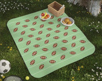 May include: A light green rectangular picnic blanket featuring a repeating pattern of brown American footballs and thin white horizontal lines. The blanket is displayed on grass with a small wooden basket and a bowl of food items.