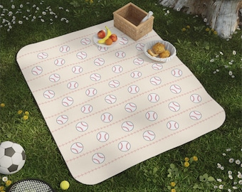 May include: A rectangular picnic blanket with a cream background and a repeating pattern of red-stitched baseballs arranged in horizontal rows. The blanket is laid out on grass with a small wooden basket, a plate of fruit, and a bowl of snacks.