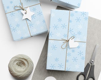 May include: Three rectangular gift boxes wrapped in light blue paper with a white and dark blue snowflake pattern. Each box is tied with brown twine and features a plain white paper gift tag in either a star or heart shape.