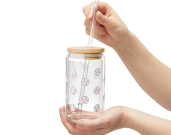 May include: A clear glass tumbler with a cylindrical shape, featuring a repeating pattern of small white baseballs with red stitching. The glass includes a light-colored wooden lid with a central hole and a straight, clear glass straw.