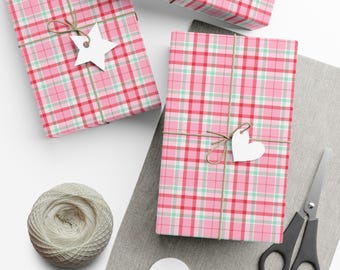 May include: Three rectangular gifts wrapped in pink, red, and mint green plaid paper, tied with brown twine. One gift features a white star-shaped tag, another a white heart-shaped tag. A ball of twine and black scissors are nearby.