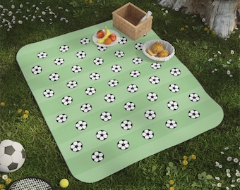 May include: A rectangular picnic blanket featuring a light green and dark green striped pattern with a repeating black and white soccer ball print. The blanket is placed on grass with a small wooden basket, a plate of fruit, and a bowl of food.