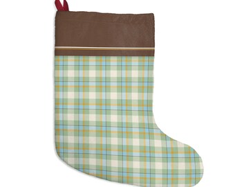 May include: A Christmas stocking featuring a brown cuff with thin white and yellow horizontal stripes. The main body of the stocking displays a plaid pattern in shades of light green, white, light blue, and yellow. A small red fabric loop is attached.