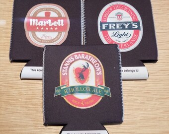May include: Three black neoprene can coolers with different designs. The first cooler features a red, white, and gold logo with the text "Martell" and "Prince of Dorne". The second cooler features a red, white, and gold logo with the text "Frey's Light" and "Lord of the Crossing". The third cooler features a red, white, and gold logo with the text "Stannis Baratheon's" and "R'hollor Ale".