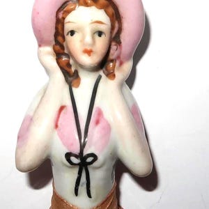 May include: A porcelain half doll figurine featuring a person with brown hair wearing a wide-brimmed pink hat. The figure has a white bodice with pink accents, a black painted ribbon tie at the chest, and a copper-colored fabric skirt base.