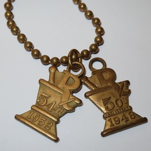 May include: A gold chain necklace with two gold charms. The charms are shaped like a mortar and pestle with the letters "Rx" on them. One charm says "54th 1952" and the other says "50th Anniversary 1948".