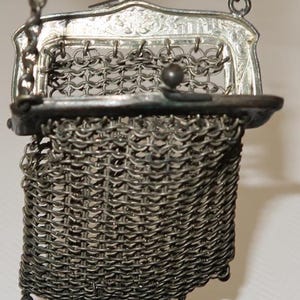 May include: A small silver-toned metal mesh purse featuring a rectangular frame with intricate engraved detailing. The bag includes a hinged clasp closure, a delicate chain handle, and three small spherical metal beads hanging from the bottom edge.