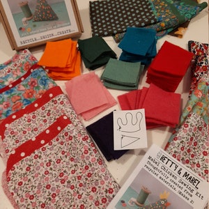 Puede incluir: Un kit de costura para hacer dos pollos de fieltro. El kit incluye una variedad de cuadrados de fieltro de colores, recortes de tela estampada, botones y un patr&oacute;n para el pollo. El kit est&aacute; etiquetado como "Betty & Mabel Mabel Chicken Sewing Kit Thoughtfully created from recycled materials. (Makes 2)" y "Recycle...Design...Create..."