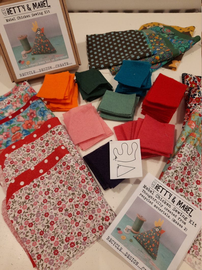 Puede incluir: Un kit de costura para hacer dos pollos de fieltro. El kit incluye una variedad de cuadrados de fieltro de colores, recortes de tela estampada, botones y un patr&oacute;n para el pollo. El kit est&aacute; etiquetado como "Betty & Mabel Mabel Chicken Sewing Kit Thoughtfully created from recycled materials. (Makes 2)" y "Recycle...Design...Create..."