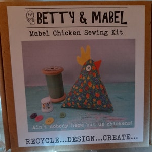 Puede incluir: Un kit de costura con un alfiletero de pollo de tela colorida, con una cresta amarilla y un pico rojo. El kit incluye recortes de tela, botones, hilo y un dedal. La caja tiene el texto "BETTY & MABEL" y "Mabel Chicken Sewing Kit" en la parte delantera. El texto "Ain't nobody here but us chickens!" y "RECYCLE...DESIGN...CREATE..." tambi&eacute;n est&aacute; en la caja.