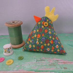Puede incluir: Un alfiletero de tela floral azul y amarilla con forma de pollo, con una cresta de fieltro amarillo y un bot&oacute;n amarillo para el ojo.