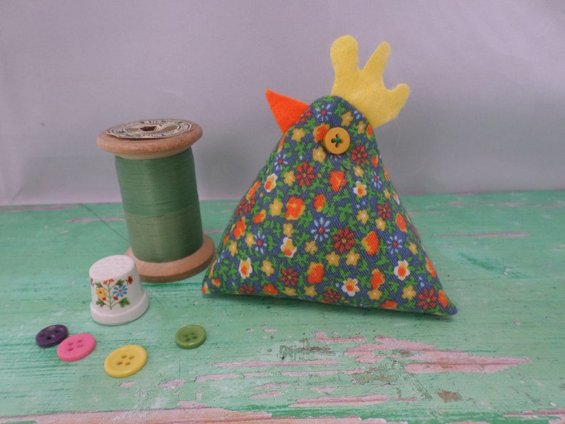 Puede incluir: Un alfiletero de tela floral azul y amarilla con forma de pollo, con una cresta de fieltro amarillo y un bot&oacute;n amarillo para el ojo.