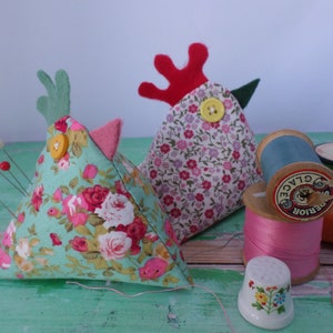 Puede incluir: Dos almohadillas para alfileres triangulares con forma de pollos, uno con cabeza verde y pico rosa, el otro con cresta roja y pico verde. Ambos est&aacute;n cubiertos de tela floral y tienen botones amarillos para los ojos. Hay carretes de hilo y un dedal en el fondo.