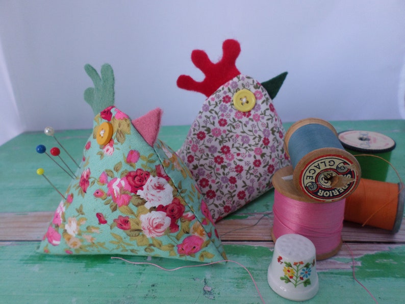 Puede incluir: Dos almohadillas para alfileres triangulares con forma de pollos, uno con cabeza verde y pico rosa, el otro con cresta roja y pico verde. Ambos est&aacute;n cubiertos de tela floral y tienen botones amarillos para los ojos. Hay carretes de hilo y un dedal en el fondo.