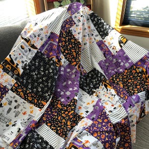 May include: A patchwork quilt with a Halloween theme. The quilt features various patterned squares in black, white, purple, and orange. Designs include skulls, pumpkins, and the words "Happy Halloween". The quilt is draped over a gray surface.