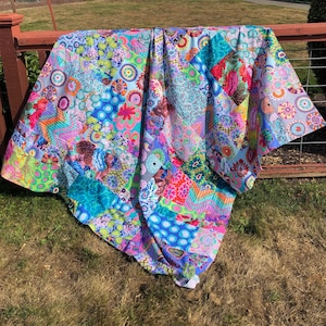 Op de afbeelding: Een kleurrijk patchwork-deken met verschillende bloem- en geometrische patronen in tinten blauw, roze, groen en paars. Het deken is gemaakt van verschillende stoffen en hangt aan een houten hek.