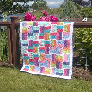 May include: A colorful patchwork quilt with a variety of bright colors, including pink, blue, green, yellow, and purple. The quilt is made of squares and rectangles, and it is hanging on a wooden fence.