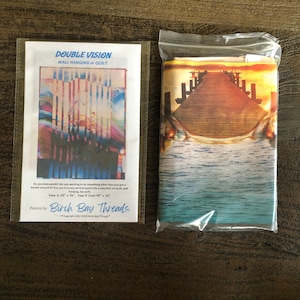 May include: A close-up of a wall hanging or quilt pattern featuring a colorful sunset over a wooden pier extending into the water. The pattern is called "Double Vision" and is by Birch Bay Threads. The pattern is available in two sizes: 39 x 56 inches and 45 x 60 inches.